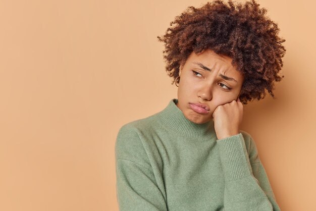 woman looks with doleful expression away leans on hand has moody look concentrated away wears casual jumper isolated on brown with copy space area