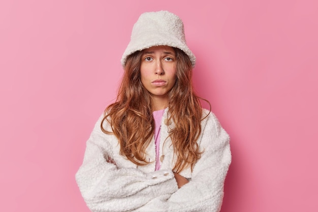 woman looks with offended expression keeps arms folded wears fur coat and hat looks hurt at unfair thing frowns and sulks poses against pink grimaces displeased