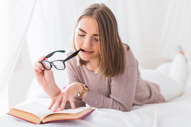 Woman lying on bed biting black eyeglasses in mouth while reading book