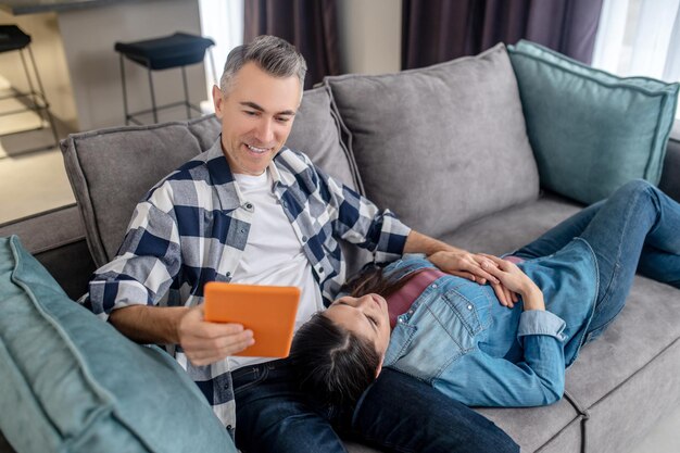 Woman lying on sofa and husband sitting with tablet