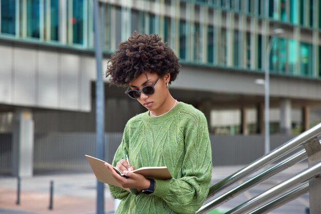 woman makes notes in notepad wears trendy sunglasses and casual green jumper poses outdoors