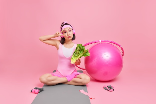 woman makes peace gesture sits crossed legs on mat holds fresh green vegetable listens music has aerobics training surrounded bu fitball hula hoop sport equipment.