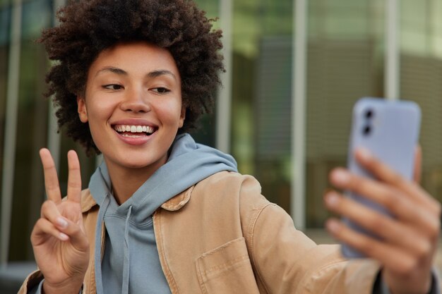 女性が平和のジェスチャーをするスマートフォンで自分撮りをするカメラが屋外でカジュアルな服を着てポーズをとるオンライン通話を行う