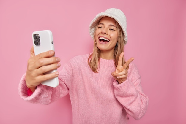 La donna fa l'autoritratto tramite il cellulare mostra il segno di pace sorride felicemente indossa un maglione casual e il cappello posa sul rosa