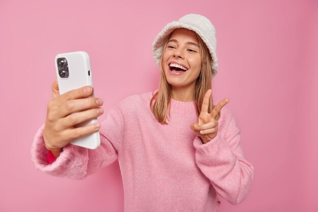 여성은 휴대폰을 통해 자화상을 만들고 평화 사인 미소를 보이며 캐주얼한 점퍼와 모자 포즈를 핑크색으로 기쁘게 입는다.