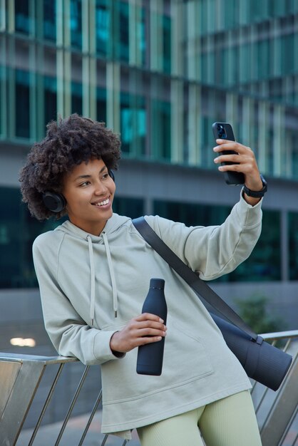 woman makes selfie satisfied after productive workout holds bottle of water dressed in sportswear carries fitness mat smiles broadly at camera poses against urban building