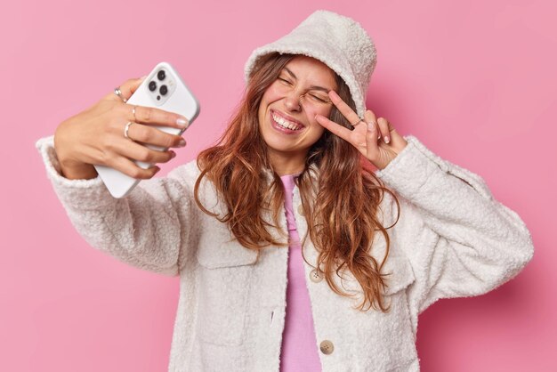 woman makes selfie on smartphone shows peace sign on eye dressed in atificial fur coat and hat enjoys video call isolated on pink smiles with white teeth.