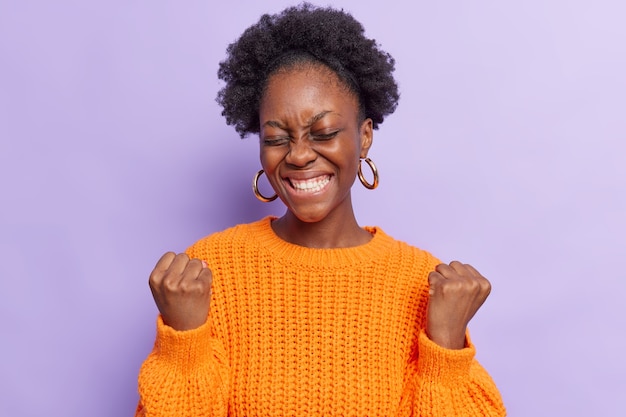  woman makes yes gesture feels like winner smiles broadly celebrates win feels excited keeps eyes closed wears casual orange knitted sweater isolated on purple 