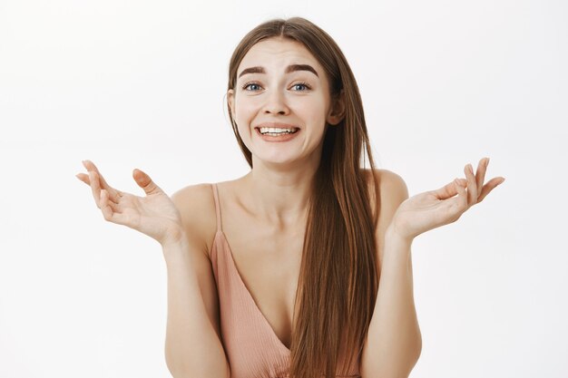 Woman making sincere surprised expression shrugging with hands spread near shoulders smiling and amazed standing impressed and astonished in beige dress