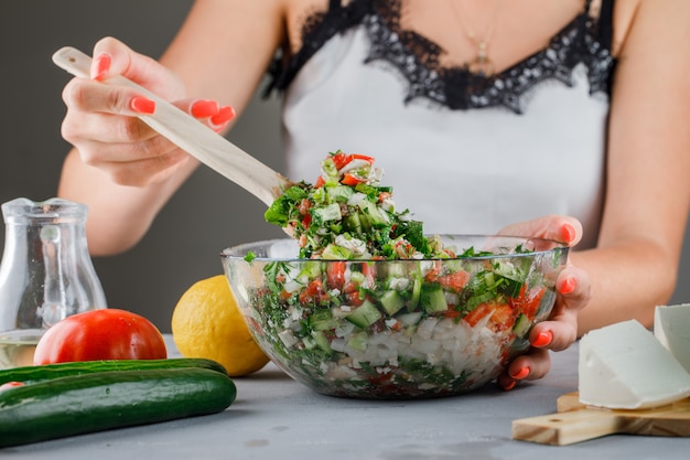 Woman making vegetable salad in glass bowl with tomatoes, cheese, cucumber on gray surface