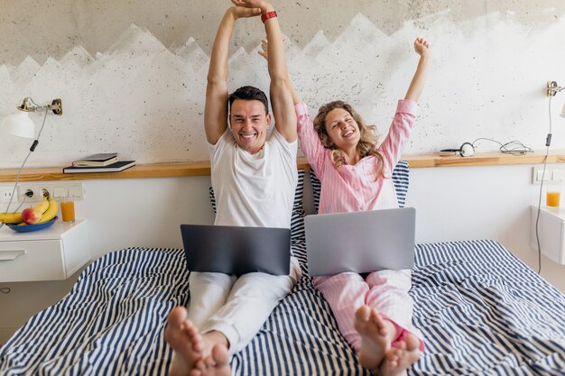 Woman and man in bed in morning smiling happy working online, family living together in bedroom wearing pajamas