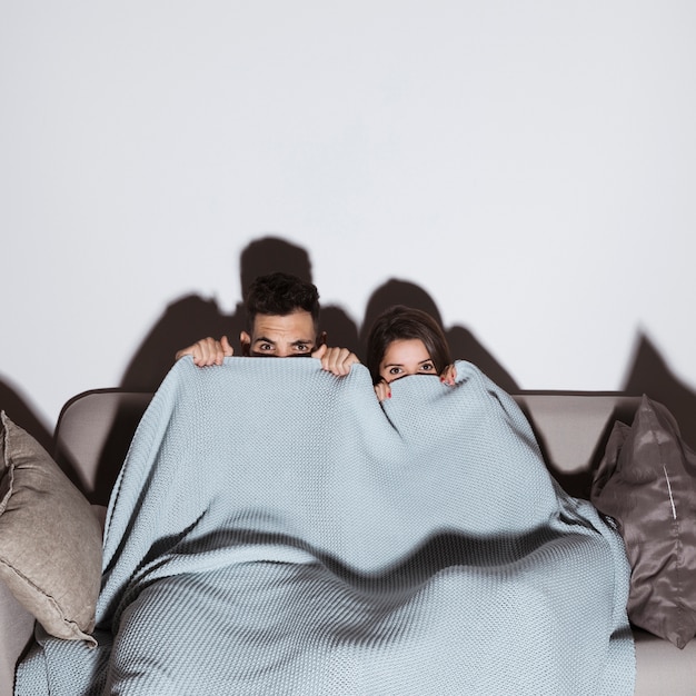 Woman and man in coverlet watching TV on sofa in dark room