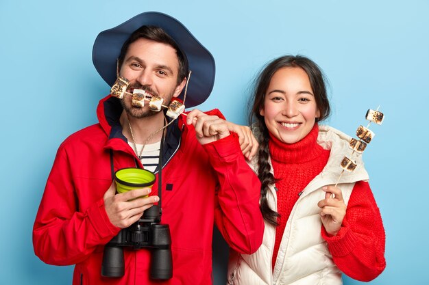 Woman and man eat roasted marshmallows made on camping fire, have picnic in forest, enjoy leisure, drink hot bevereage, wears casual outfit, pose against blue wall