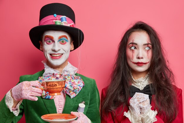 Woman and man in halloween costumes and professional makeup pose indoor against pink wall. Mad hatter from wonderland drinks tea