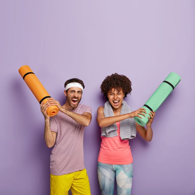 Woman and man hold rolled up karemats, shout loudly, dressed in casual clothes, have yoga training