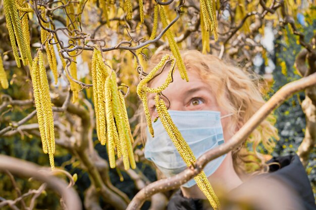 マスクをした女性が開花ヘーゼルに触れる花粉症の概念春のアレルギーの主な引き金開花ヘーゼルの尾状花序に選択的に焦点を当てる