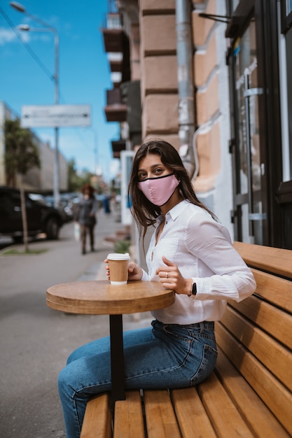Woman in a medical mask drinks coffee on the street