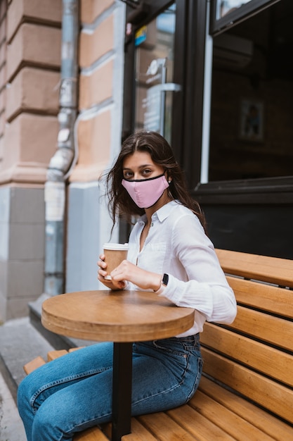 바이러스 감염을 예방하기 위해 의료 마스크를 쓴 여성이 길거리에서 커피를 마신다