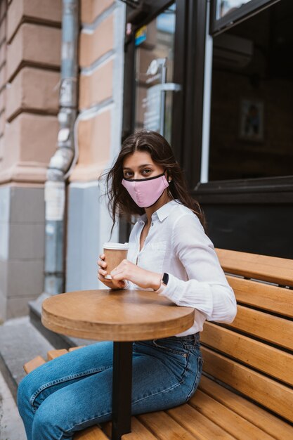 Woman in a medical mask in order to prevent viral infections drinks coffee on the street