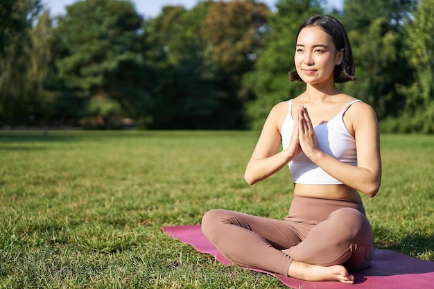 Женщина медитирует на лужайке в парке, сидя на спортивном коврике, расслабляясь, дыша свежим воздухом