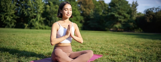 スポーツマットの上に座って公園の芝生で瞑想し、新鮮な空気を呼吸してリラックスする女性