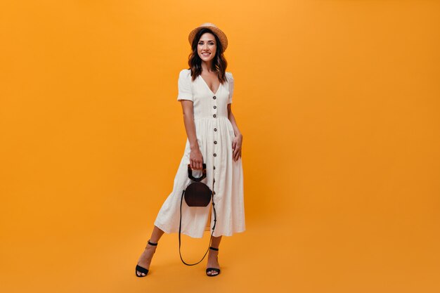 Woman in midi dress and straw hat posing with bag on orange background