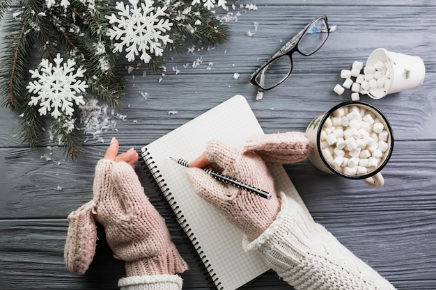 Woman in mittens writing in notebook near cup with marshmallow and eyeglasses 