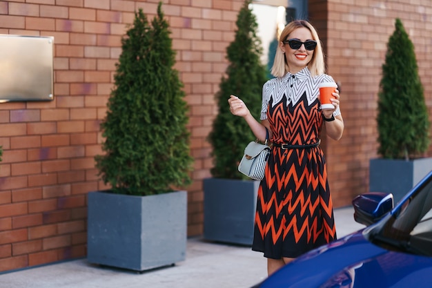 Foto gratuita donna in movimento con un caffè per andare mentre si cammina vicino a un'auto in città