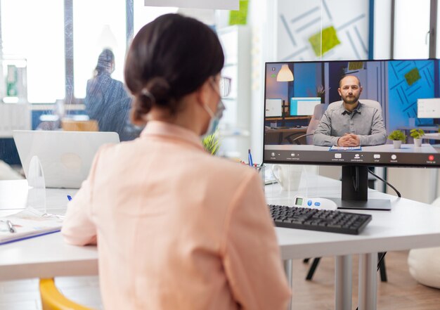 Woman in new normal office listening man talking during online video conference, looking at screen discussing project during coronavirus flu outbreak.