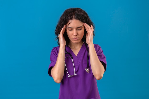 woman nurse in medical uniform and with stethoscope feeling fatigue touching temples having headache standing on isolated blue