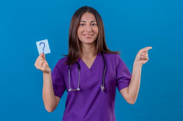 의료 제복을 입은 여자 간호사와 청진기는 파란색에 서있는 측면에 손과 손가락으로 매우 행복하게 가리키는 물음표로 알림 용지를 들고