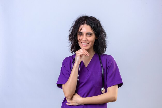 woman nurse in medical uniform and with stethoscope with hand on chin smiling confident standing on isolated white