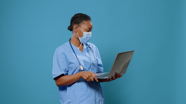 여성 간호사는 얼굴 마스크를 쓰고 카메라 앞에서 노트북을 사용합니다. 제복을 입은 의료 조수는 코로나바이러스로부터 보호하면서 건강 관리를 위해 컴퓨터를 들고 있습니다.