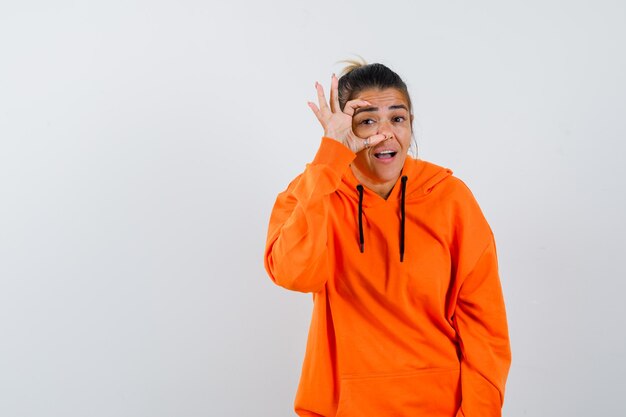 Woman in orange hoodie opening eyes with fingers and looking curious