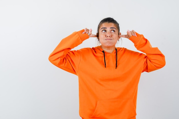 Woman in orange hoodie plugging ears with fingers and looking curious