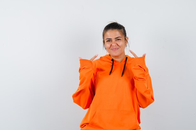 Woman in orange hoodie pointing at herself and looking happy