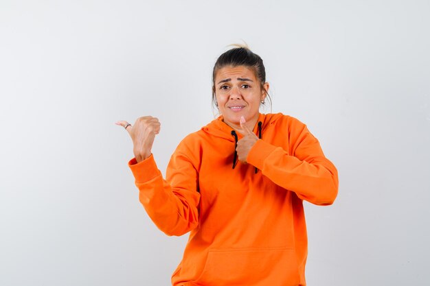 Woman in orange hoodie showing thumb up, pointing aside and looking confused