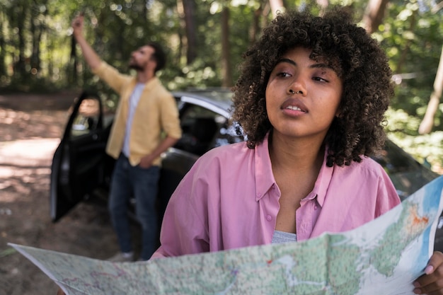 Woman outdoors holding map while boyfriend takes selfie next to car