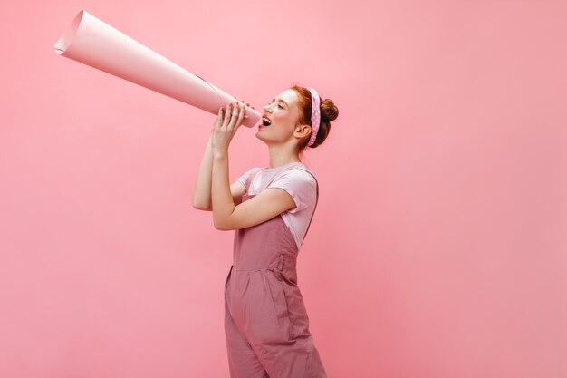 Free photo woman in overalls shouts into horn and looks into camera on pink background.