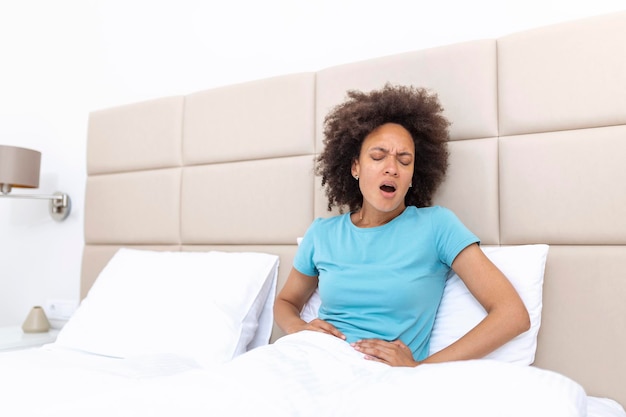 Woman in painful expression holding hands against belly suffering menstrual period pain lying sad on home bed having tummy cramp in female health concept