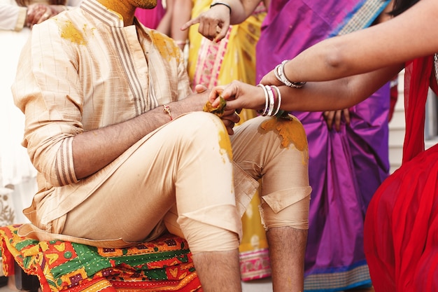 Woman paints Indian groom's knees and face with turmeric paste