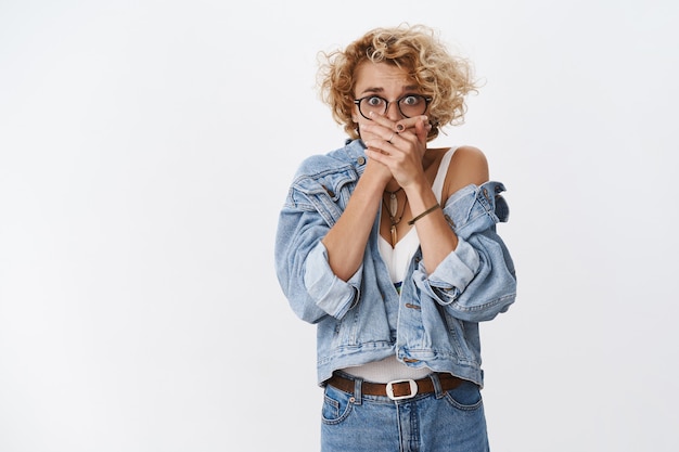 Woman in panic witnessing terrible crime shut mouth with palms nervously not scream freak out, popping eyes at front horrified and shocked standing speechless over white wall