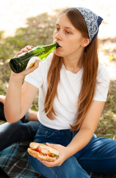 Женщина в парке пьет пиво и ест гамбургер