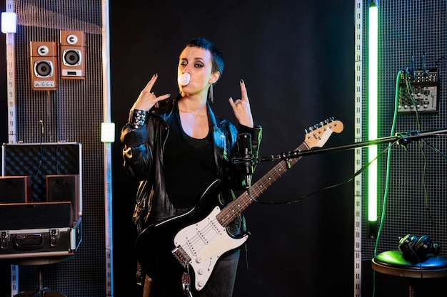 Woman performer blowing bubble gum while posing in studio doing rockstar gesture, preparing electric guitar before performing metal concert. Rebel musician playing for solo performance