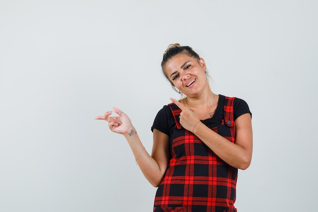 Woman in pinafore dress pointing aside and looking merry , front view.