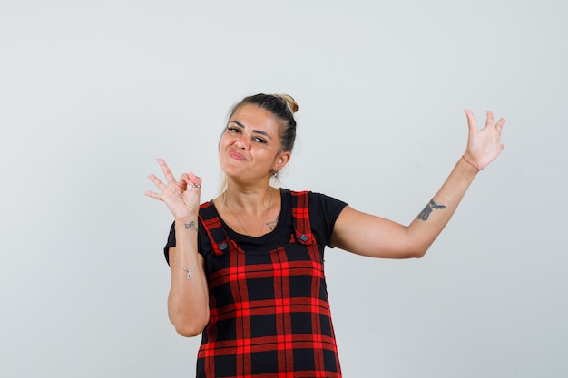 Woman in pinafore dress showing ok gesture, raising arm and looking confident , front view.