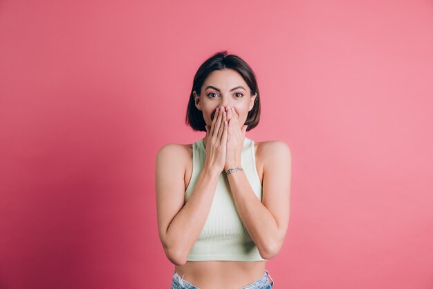 Woman on pink background covering mouth with hands looking at camera, funny excited with unexpected positive surprise