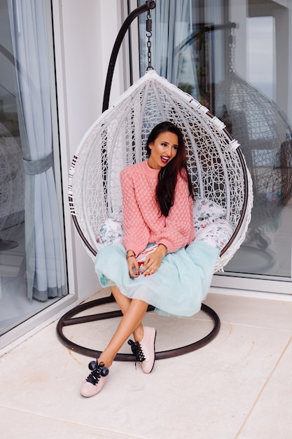 Woman in pink sweater and blue skirt stand by egg chair holding candy box