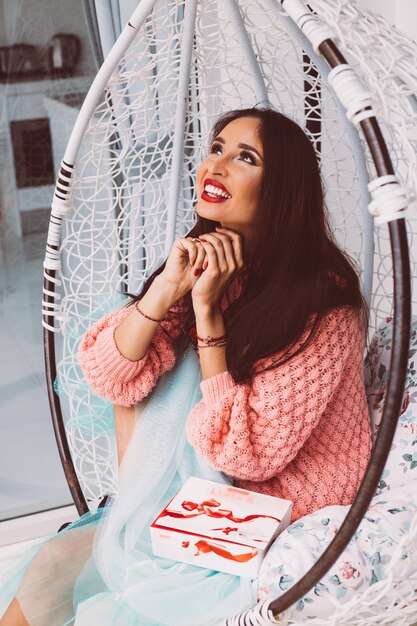 Woman in pink sweater and blue skirt stand by egg chair holding candy box