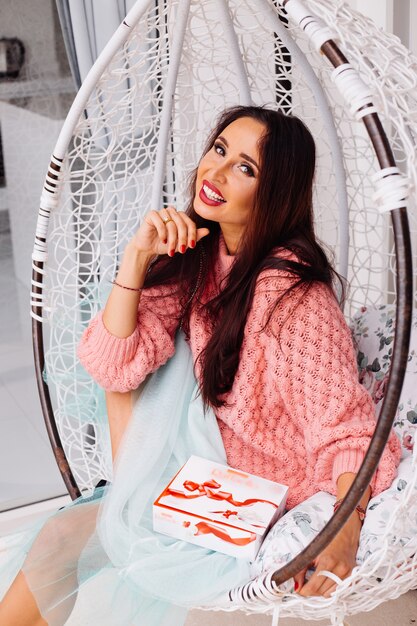 Woman in pink sweater and blue skirt stand by egg chair holding candy box